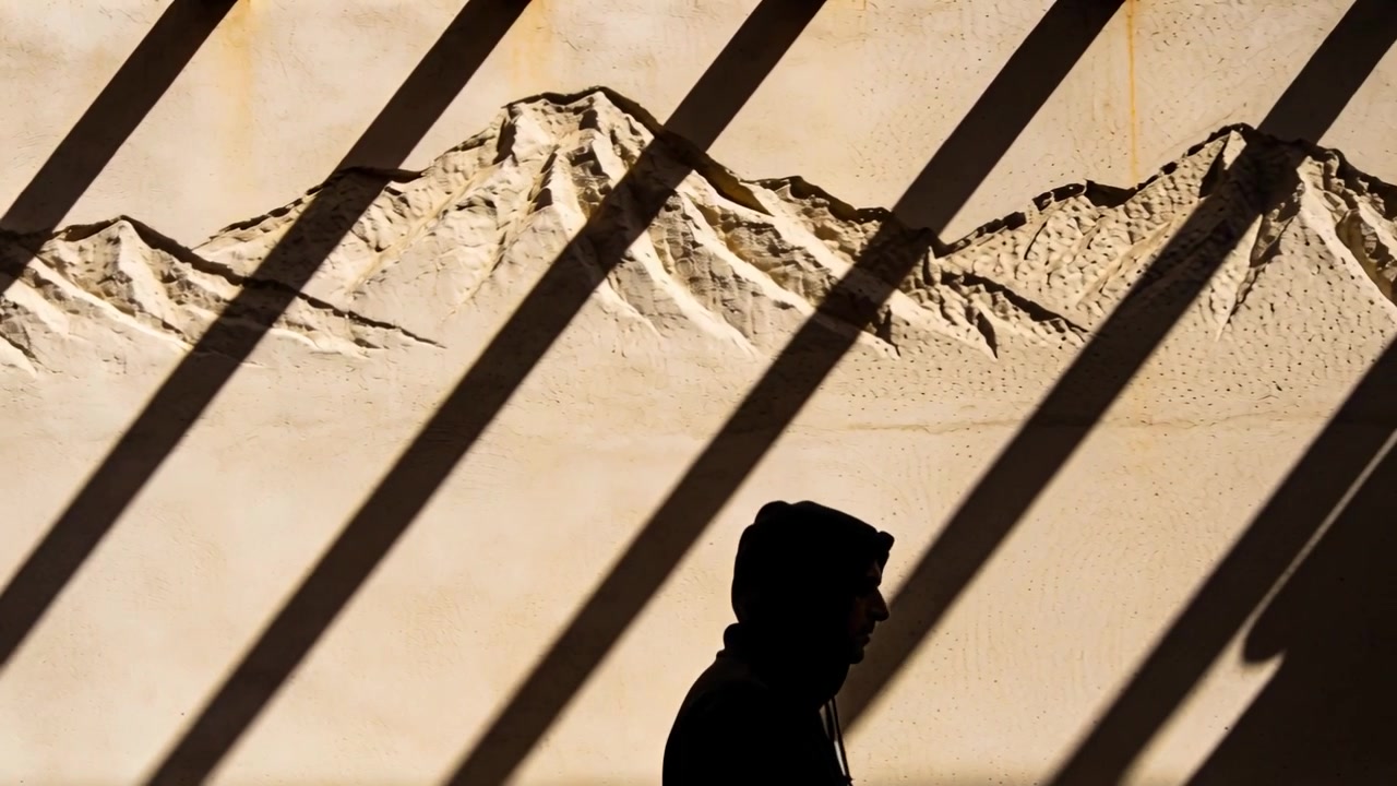 Homme à capuche entrant par la gauche, marchant vers la droite le long d'un chemin devant une fresque murale de montagne peinte avec des ombres obliques.