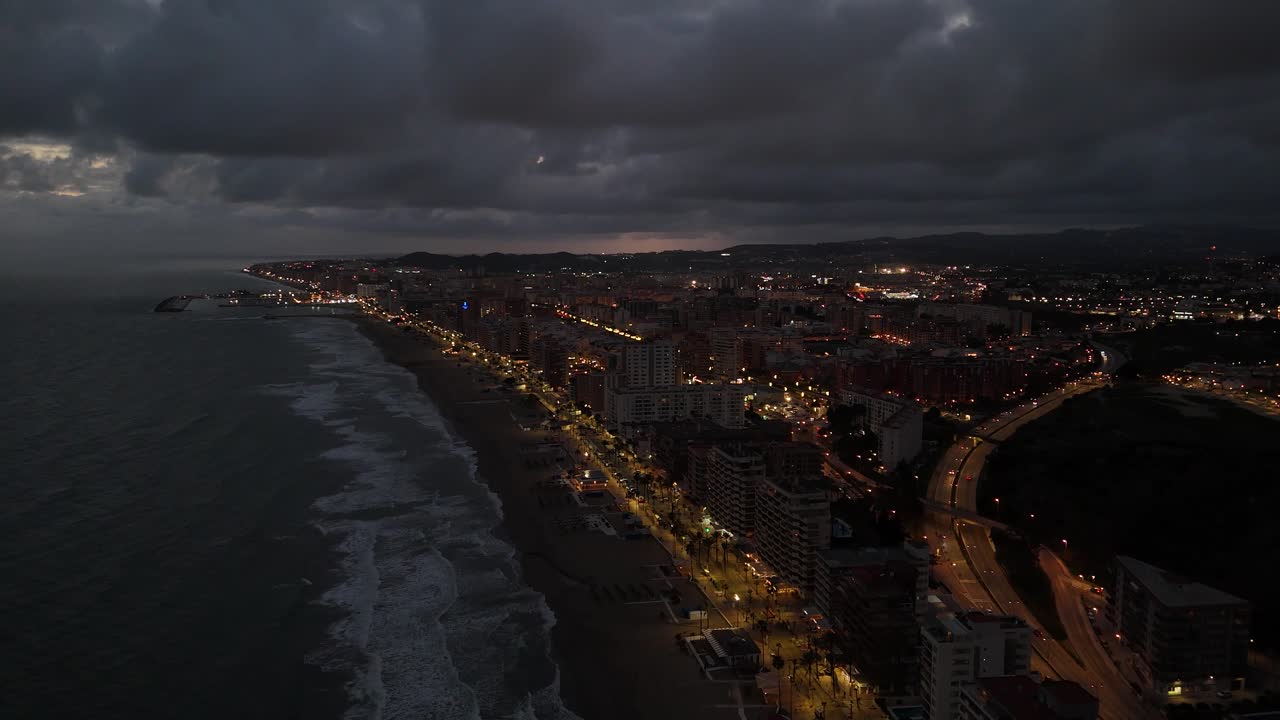 Torreblanca, Spain is glowing with lights at night, viewed from a high vantage point that showcases the coastline, buildings, a cloudy sky adding a touch of drama to the scene, drone pulling out shot