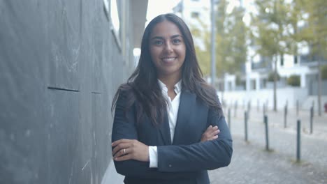 feliz empresaria latina de pie con los brazos cruzados cerca del edificio de oficinas, posando y sonriendo a la cámara