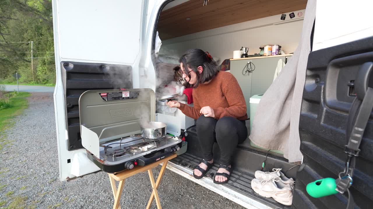 Donna che prepara un pasto su un fornello da campeggio mentre il suo cane osserva dall'interno del camper, godendosi la vita all'aria aperta.