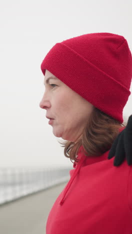 side view of athlete in red hoodie performing shoulder stretch by turning arm outdoors in snowy park, background features frosted tree, bench, snow-covered landscape, and misty atmosphere
