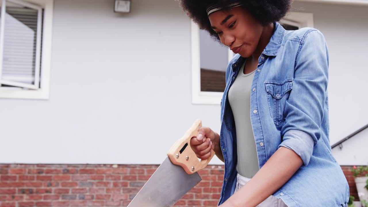 African American woman sawing wooden plank at porch workbench using hand saw and safety glasses