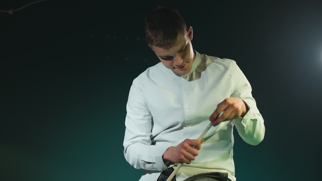 joven con camisa blanca contra un fondo oscuro sostiene cuidadosamente el palo de la pilla, aplicando la cubierta roja con expresión enfocada. la iluminación destaca la preparación, la concentración y la precisión en el billar