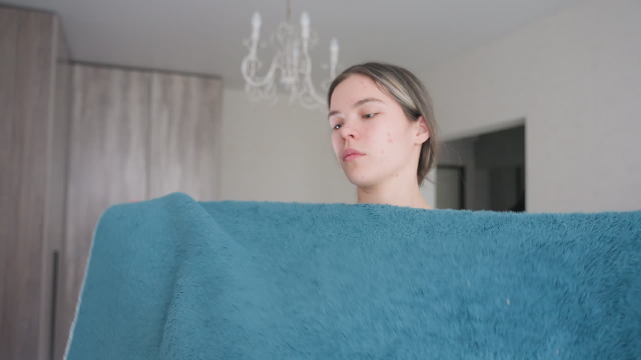 Young woman folding fluffy blue blanket indoors, hands carefully arranging soft fabric with focused expression, natural daylight brightening home setting