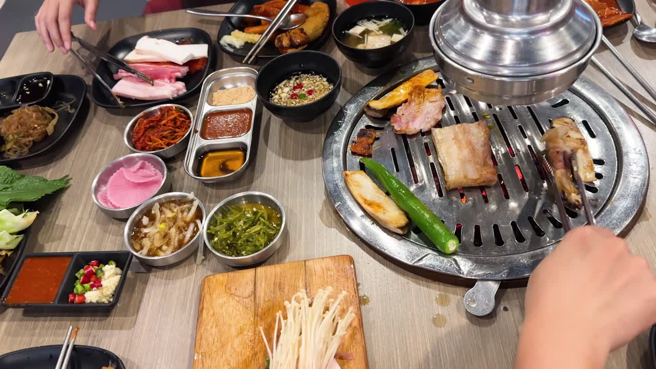 Hands grill pork and vegetables on a tabletop BBQ in a vibrant Bangkok setting. Bright lighting enhances the colorful array of side dishes