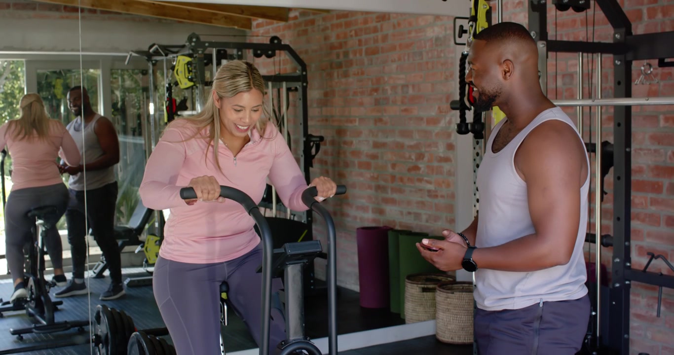 After gripping handles pink-top client pedaling air bike at gym, trainer checking phone for metrics