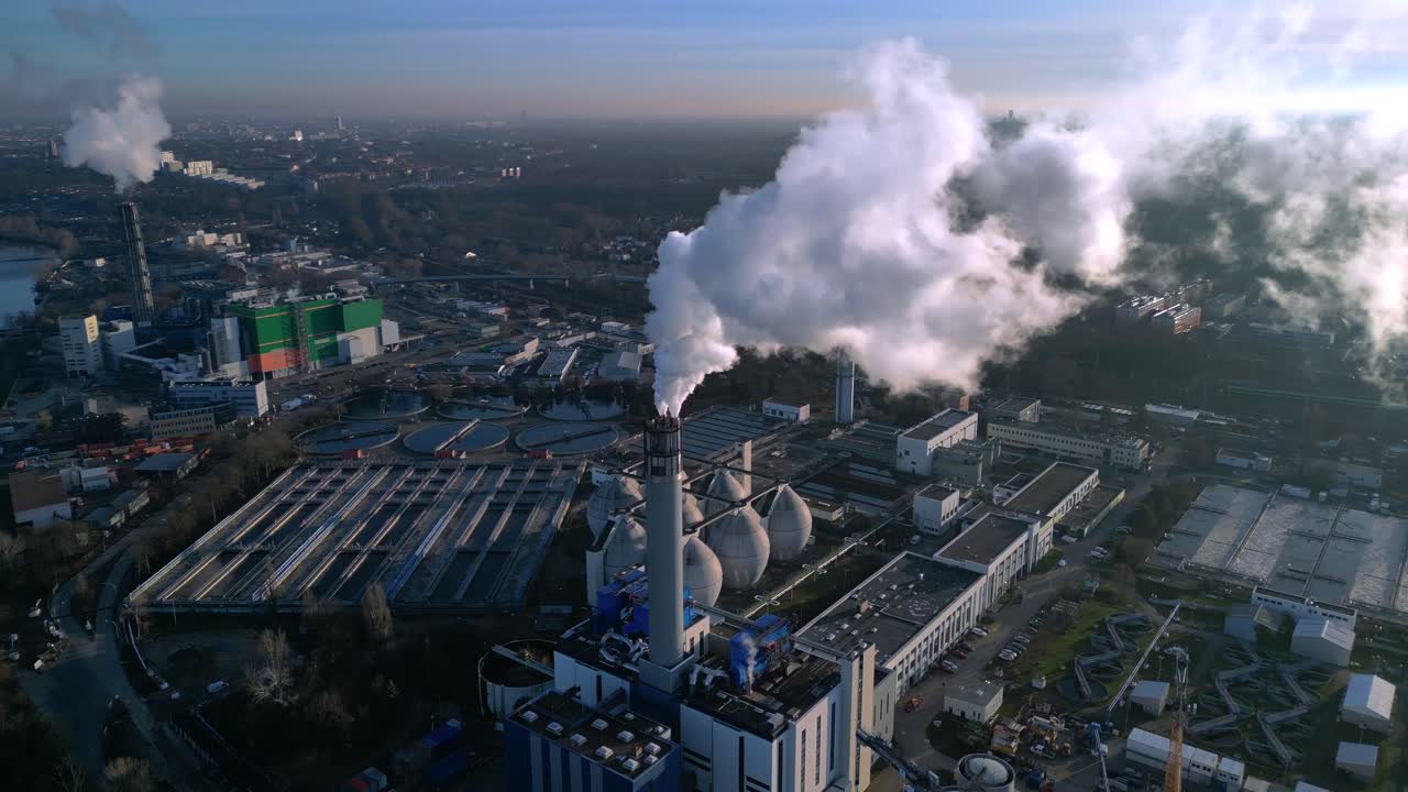 Complesso industriale con una ciminiera che emette fumo nel cielo, vicino a un impianto di trattamento delle acque reflue e infrastrutture urbane. Speed ramp hyper motion time lapse. Ottima ripresa aerea circolare con drone