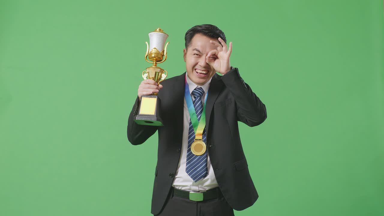 슈트와 타이를 입은 아시아 사업가, 금메달과 트로피를 들고, 오케이, 손으로 눈을 감고 카메라에 미소 짓고, 스튜디오의 녹색 스크린 배경에서 첫 번째 우승자로