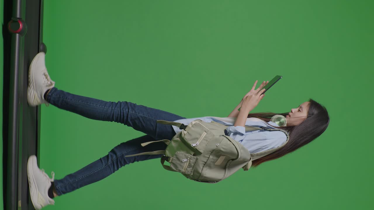 cuerpo lleno de vista lateral estudiante adolescente asiática con una mochila sonriendo y usando una tableta mientras camina en la cinta de correr en el estudio de fondo de pantalla verde