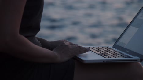 Girl-hands-with-laptop-sitting-on-rock-by-the-sea-on-sunset,-slow-motion,-close-up