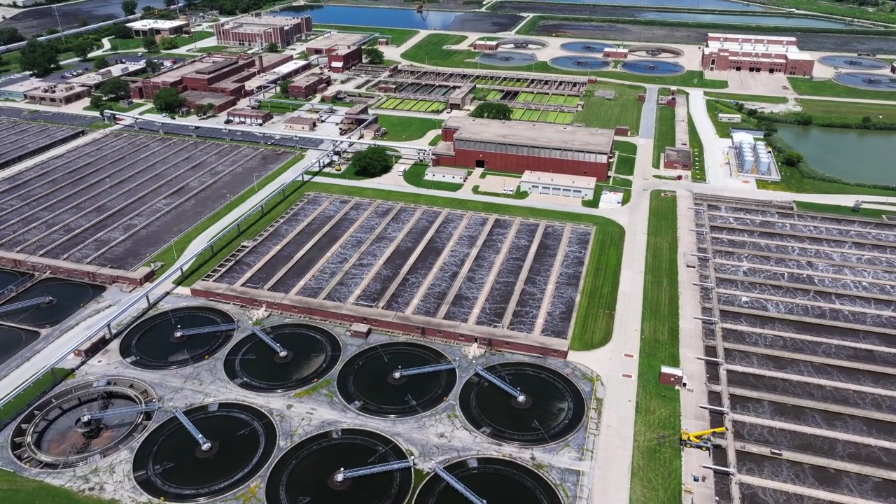 Forward drone shot above a large water reclamation facility revealing circular clarifiers, aeration channels, brick operations buildings and settling ponds beyond.