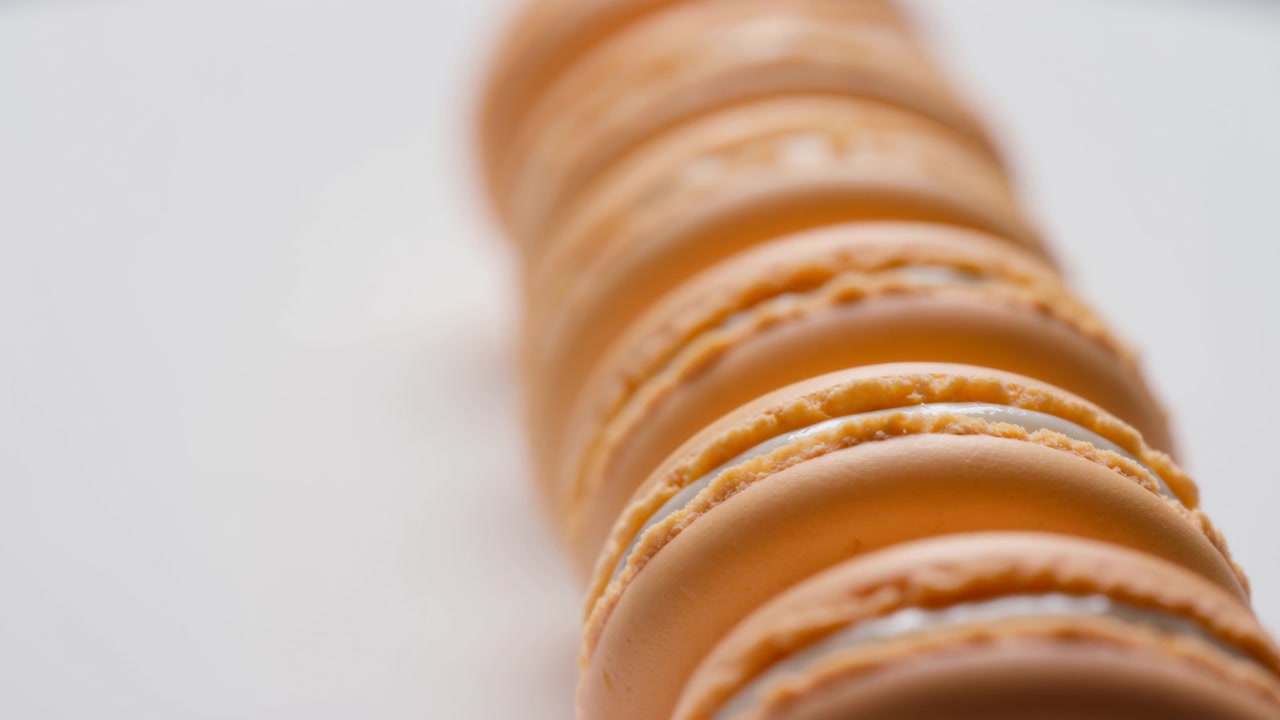 Primo piano estremo che mostra macaron color pesca-arancio impilati e allineati diagonalmente con gusci lisci a cupola, delicati piedini arricciati, ripieno pallido e luce laterale soffusa e diffusa su uno sfondo neutro e pulito.