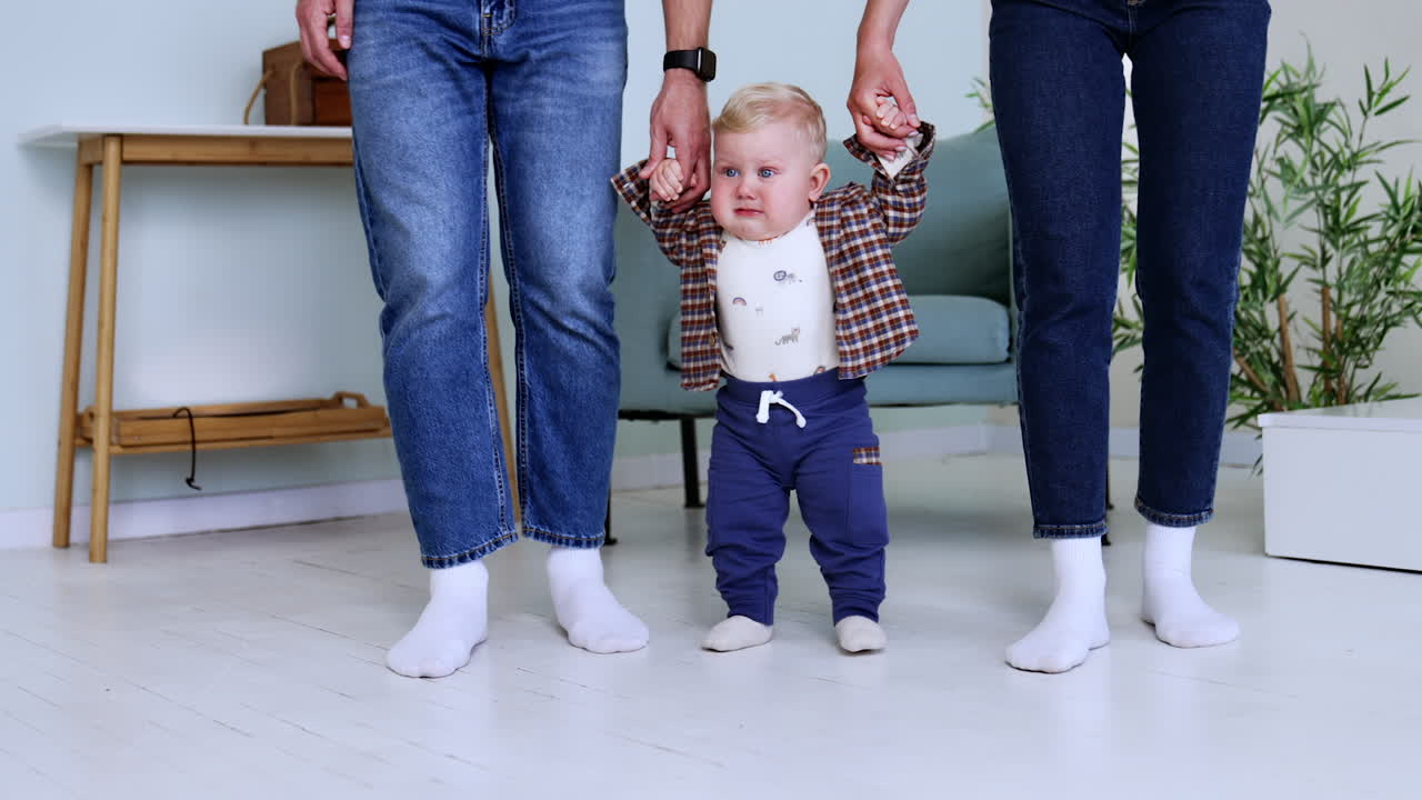 Sweet cute blond baby boy taking his first steps. Unrecognized parents support their lovely son.