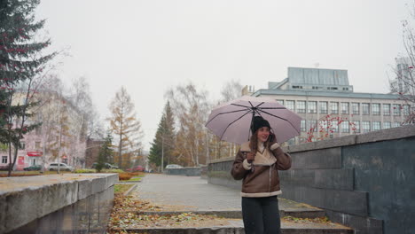 dreamy girl smiling on phone call holding umbrella, wearing knit cap, brown shearling jacket, black trousers, walking down stone steps during light snowfall with autumn leaves