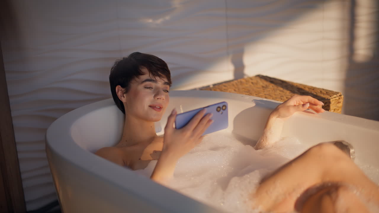 Bathing girl watching smartphone resting online in bubble foam bathtub closeup
