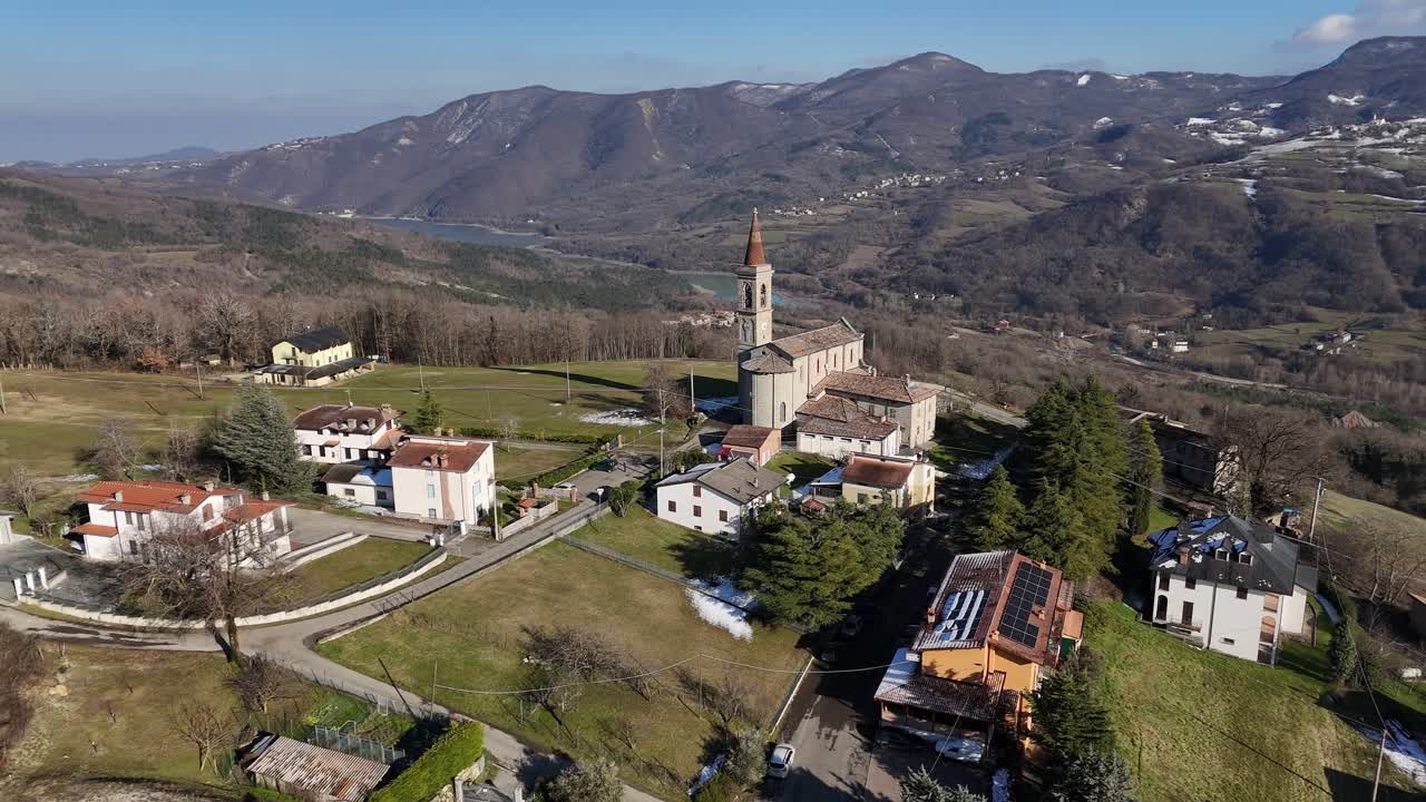 静かなI Rabbiniの上にそびえ立つSanti Salvatore e Galloの鐘楼。澄み切った晴天の下、緑のアペニン山脈の谷間にたたずむ伝統的な石造りの教会と家々。ドローンから見たイタリア、Morfasso PC.