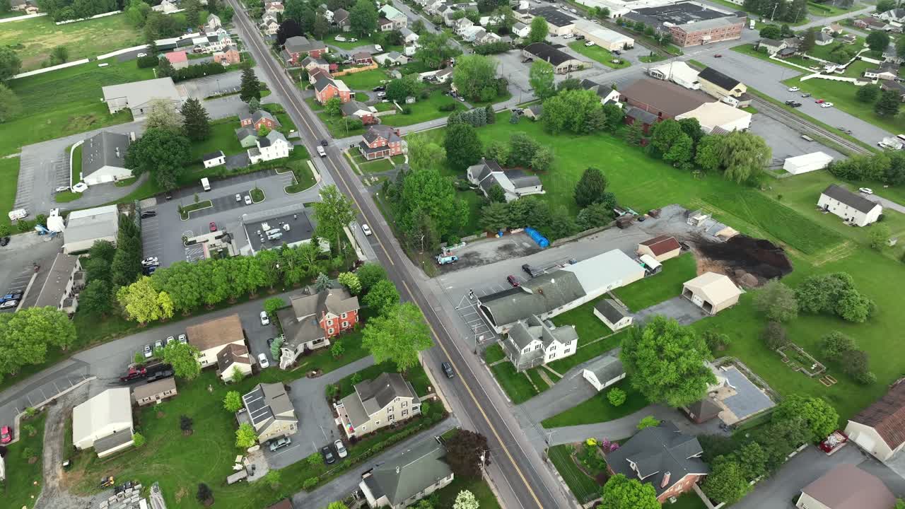 Slow drone flight over small city with traffic on main street.Idyllic green farm fields and trees in spring. Top down shot. Cloudy day in United States. Homes and houses with warehouses.