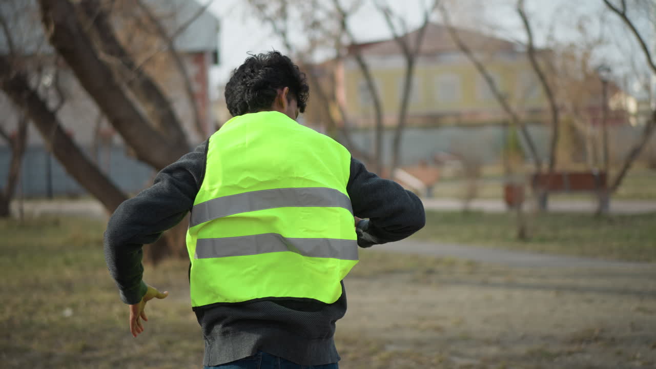 Man outdoors putting on bright neon green safety vest while standing in park with leafless trees, preparing for outdoor cleanup or volunteer work, backpack with gloves and water bottle placed on grass