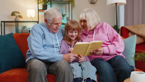 child girl granddaughter with grandparents reading interesting book together sitting on home couch