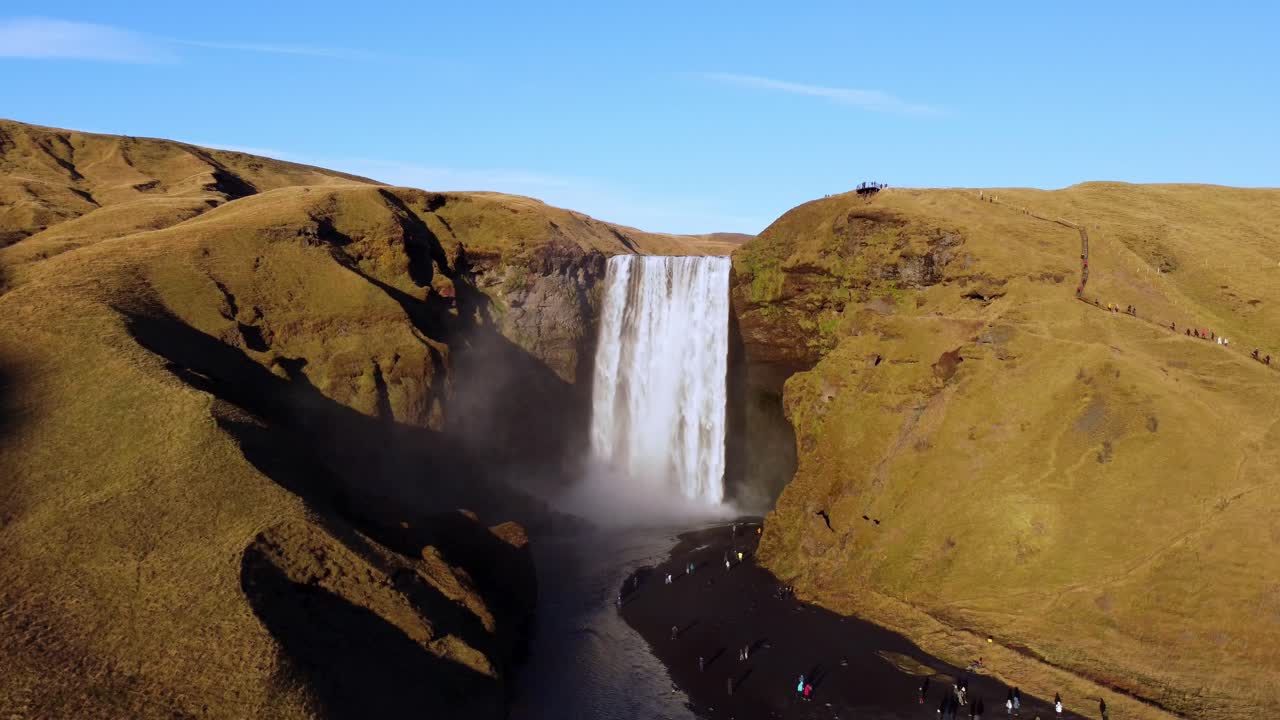 A breathtaking view of the powerful Skógafoss waterfall plummeting into a riverbed, framed by rolling, golden-brown hills under a clear blue sky in South Iceland