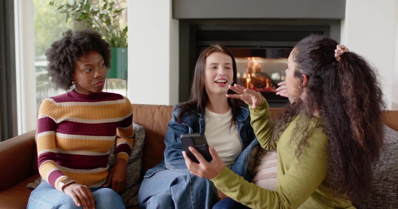 Female friends sitting on couch, rightmost woman holding smartphone sharing clip, others laughing