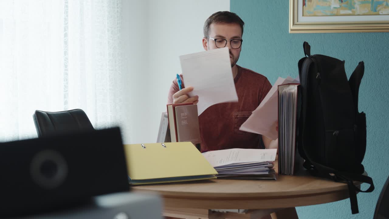 A man sits at his home desk, holding multiple sheets and marking them with a pen—focused on reviewing or correcting documents, possibly exams, legal files, or financial papers