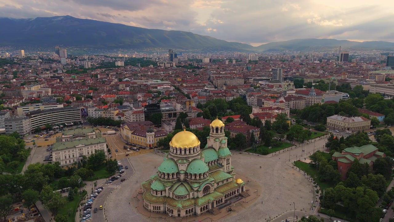 Perspectiva de drone deslizando pelas cúpulas douradas cintilantes da Catedral Alexander Nevsky ao pôr do sol, exibindo o extenso horizonte de Sófia, capital da Bulgária.