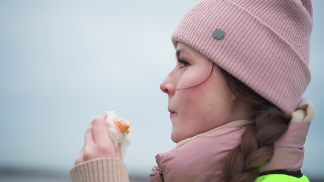 Young female worker in pink knit hat and braided hair wearing warm jacket and neon reflective vest, standing outdoors in cold weather, looking focused and thoughtful during work in urban