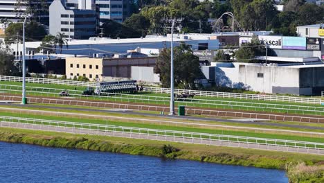a group of jockeys ride thoroughbred horses at speed along a sunlit turf racetrack, with urban gold coast buildings in the background and steady camera panning