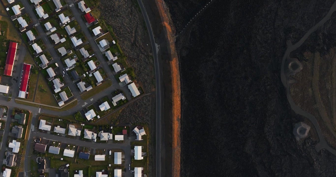 Top-down drone view of Grindavik, Iceland, with dense housing blocks beside a road and orange defense berm sharply dividing the town from an immense hardened black lava field.