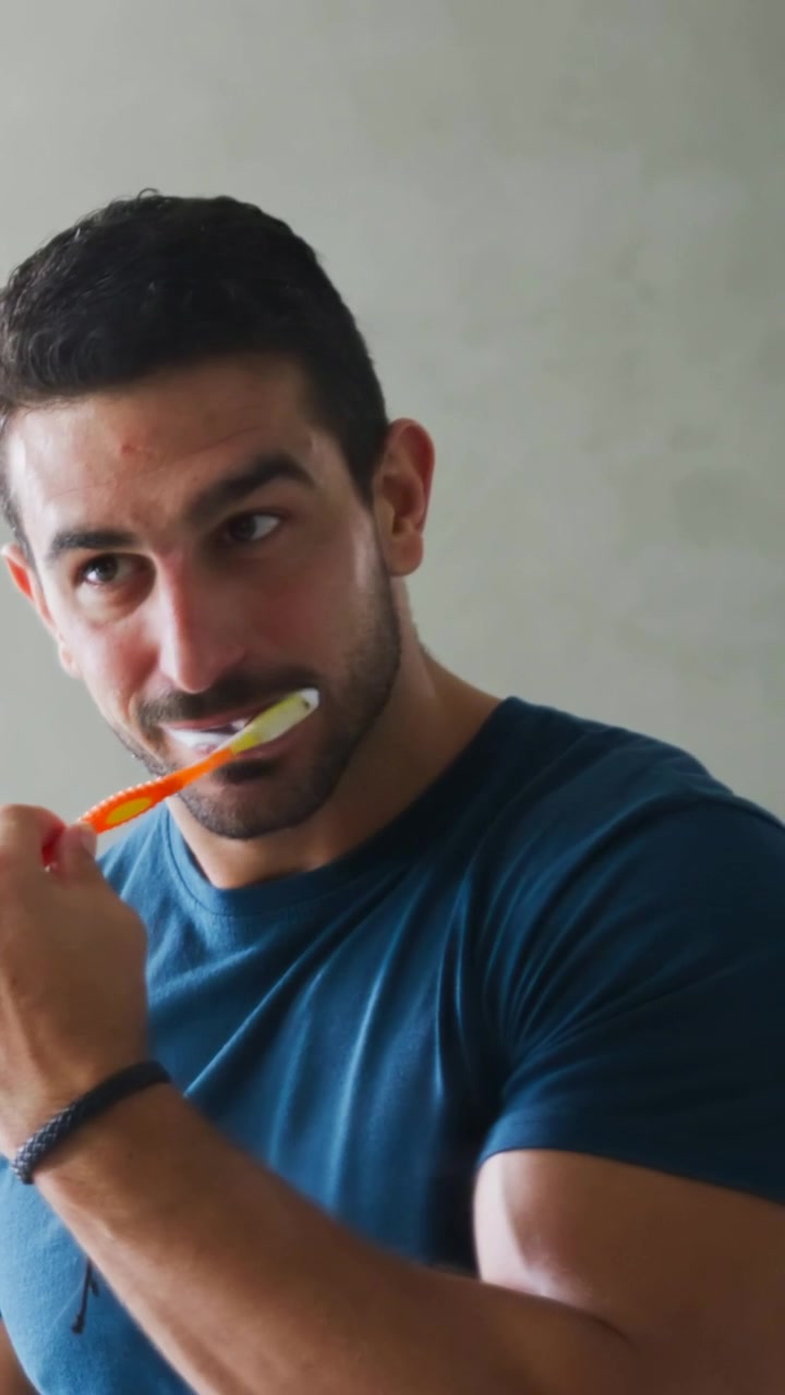 Vertical video: Raising orange toothbrush, man brushing teeth at home in navy tee and dark bracelet