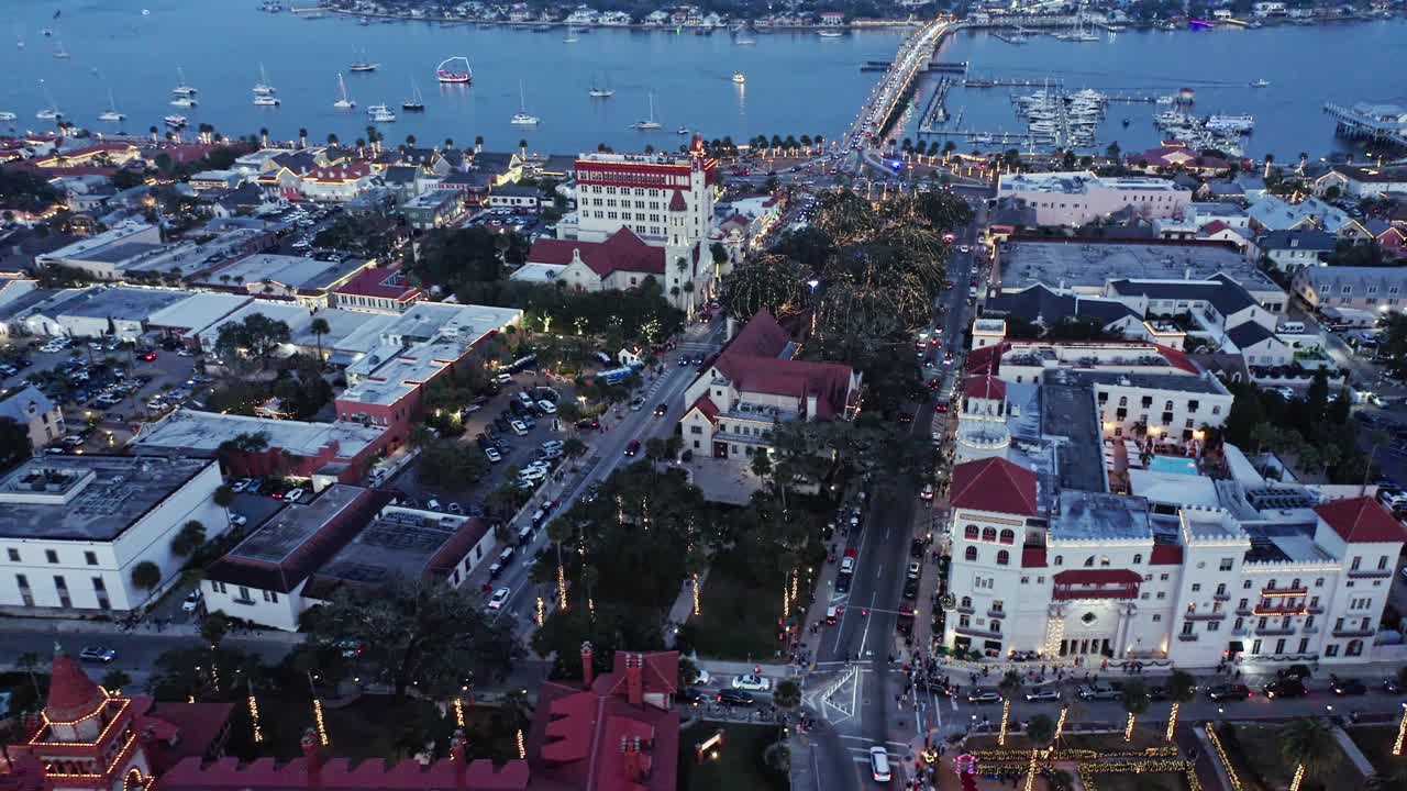 Centro histórico de la ciudad de San Agustín con edificios de estilo español, palmeras, calles concurridas y el Puente de los Leones sobre la bahía; las luces de la ciudad al anochecer comienzan a iluminar la zona