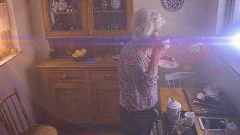 Animation-of-light-moving-over-senior-caucasian-woman-in-kitchen-looking-out-of-window-drinking-tea