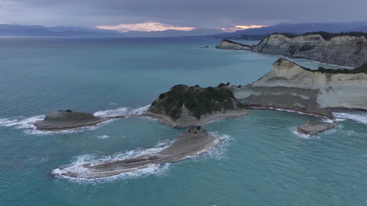 Vista aerea mozzafiato di Capo Drastis a Corfù, Grecia. Acque turchesi si infrangono contro imponenti scogliere di argilla bianca sotto un cielo al tramonto suggestivo. Pura serenità naturale.