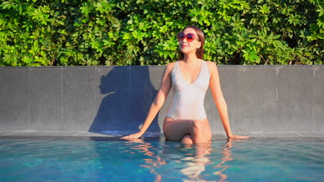 while sitting on the edge of a swimming pool, a pretty woman in a bathing suit and sunglasses looks around at her surroundings