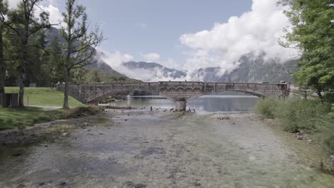 Frontales-Drohnenvideo-über-Dem-Bohinj-See,-Das-Sich-Auf-Die-Meisten-Brücken-Zubewegt,-Die-über-Den-Flussspiegel-Fliegen,-Ein-Kanu-Und-Radfahrer-Sind-Zu-Sehen,-Die-Die-Brücke-Passieren