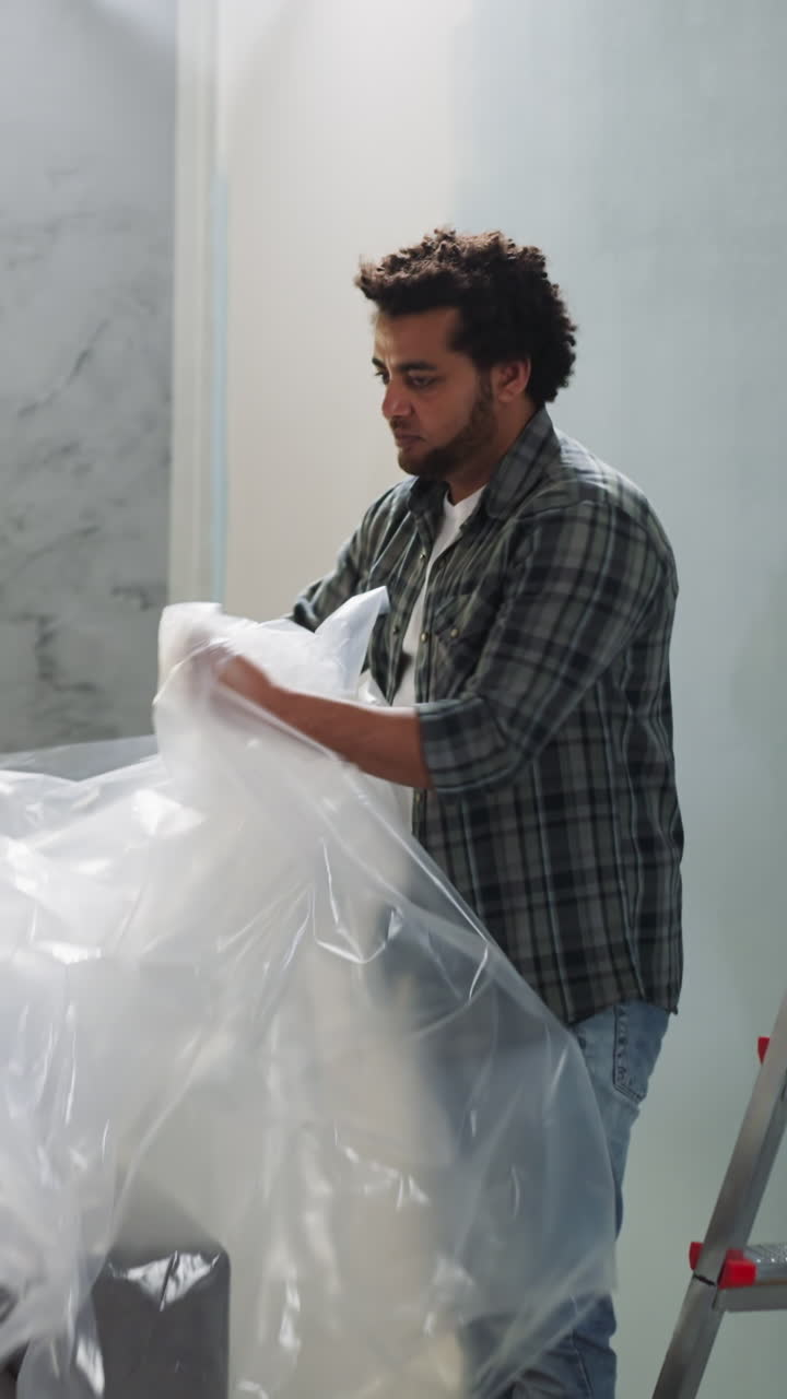 Bearded black man takes off plastic sheet from sofa in living room. African-American builder finishes room renovation. Home reconstruction process