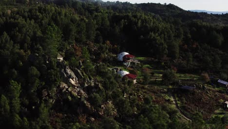 Drone-shot-of-sun-drenched-hilly-landscape-with-rolling-mist,-forest-and-small-buildings