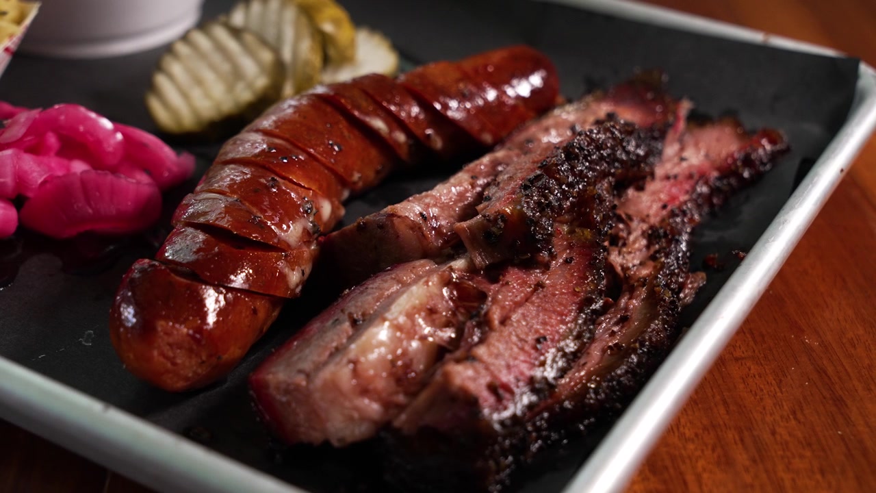 Invitante piatto di barbecue texano con due tipi di carne, con tenero brisket a fette, salsiccia affumicata, cetriolini e cipolle sottaceto su un vassoio di metallo sotto una luce calda, primo piano, slider, 4K