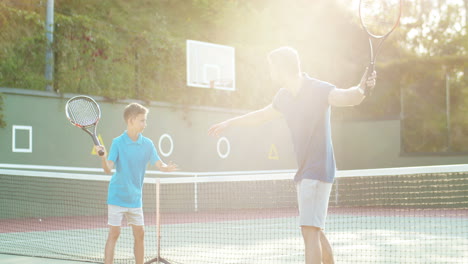 man teaching his teen son how to play tennis on an outdoor court in summer and then giving high five