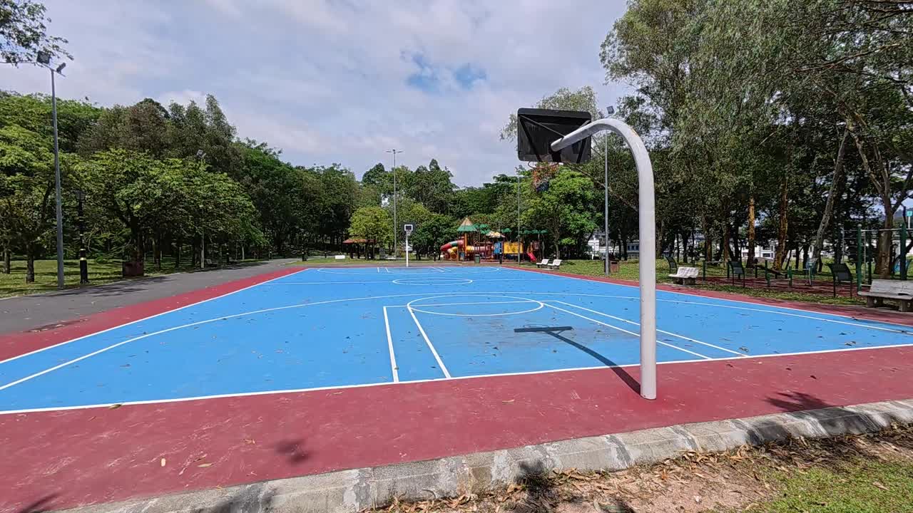 An empty basketball court in a tropical park, surrounded by lush green trees under a bright, sunny sky. Perfect weather.