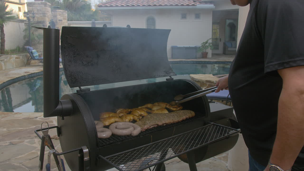 Uomo apre un barbecue fumante e sposta pollo e salsiccia sulla griglia.