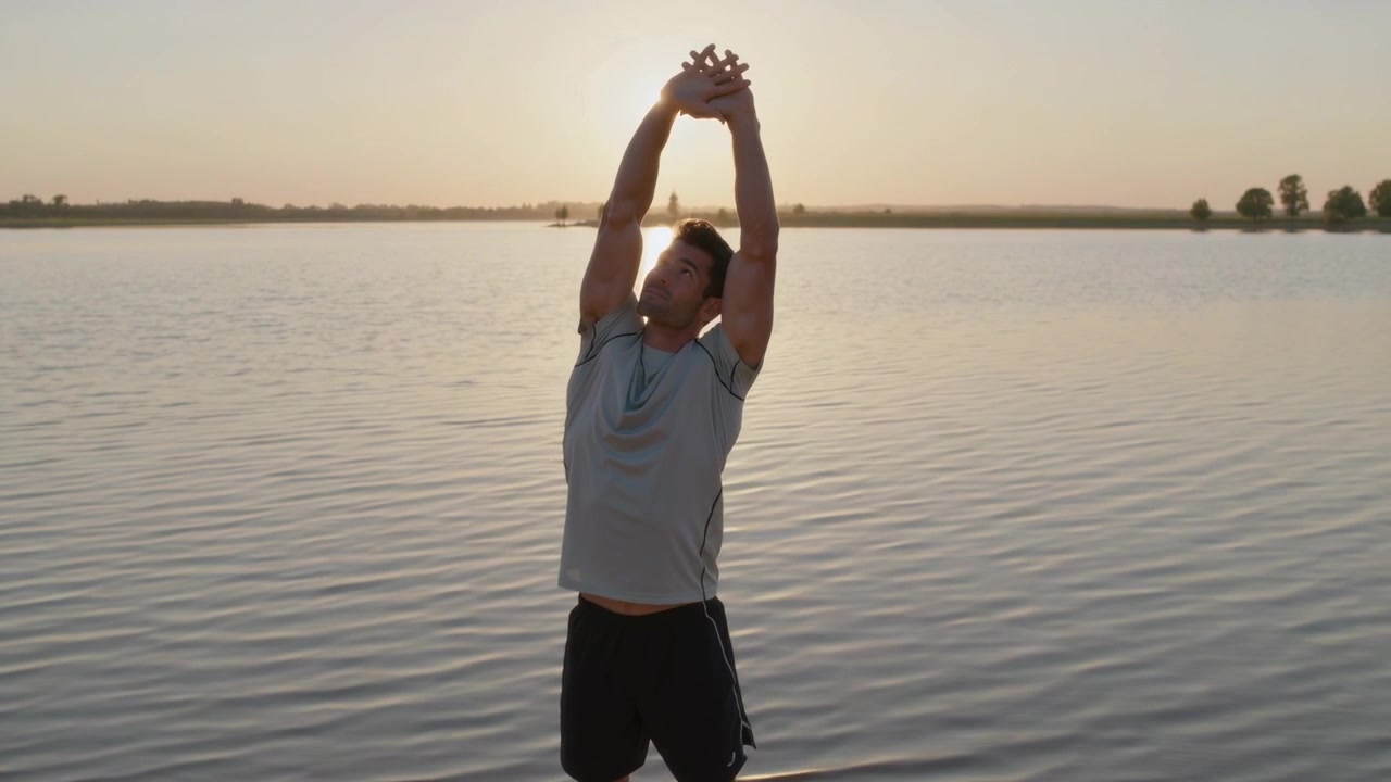 Uomo che si allunga con le mani unite, in abbigliamento sportivo, appoggiato a destra sulla sabbia in riva al lago, mentre si riscalda all'alba.