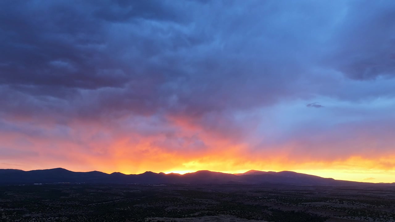 4K UHD drone timelapse: massive heavy storm cloud system dominates skyscape. Intense orange sunset colors glow against dark mountain silhouettes and sweeping scrubland mesa terrain.