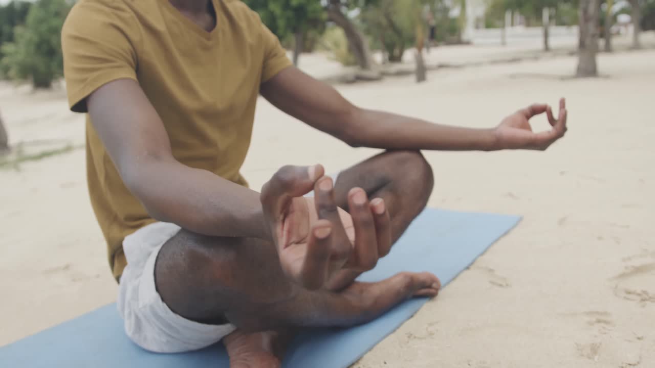 sección baja de hombre afroamericano haciendo yoga, sentado en una alfombra meditando en la playa, cámara lenta.