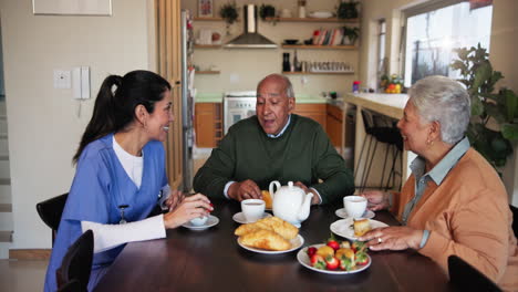 una enfermera sonríe mientras conversa con una pareja de ancianos mientras beben té y pasteles.