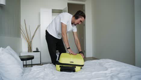 happy bearded man getting ready for holiday putting in clothes in suitcase at home background. handsome man in black headphones