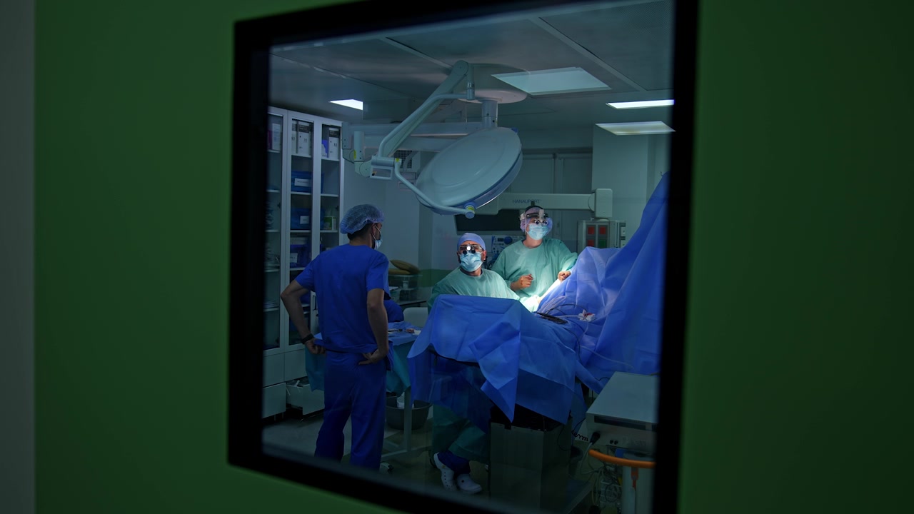 Surgeons in surgical loupes perform operation on trauma. Doctors show thumbs up to the camera through the window in the door. Male medic in blue robe comes up to operating table.