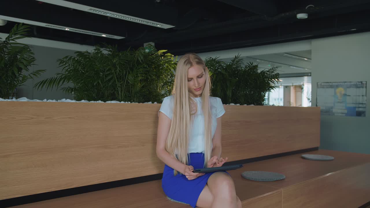 사무실에서 태블릿을 가지고 휴식을 취하는 여성. 현대적인 사무실에서 나무 트리에 맨발로 앉아 태블을 사용하고 휴식을 취하고 있는 젊은 우아한 여성