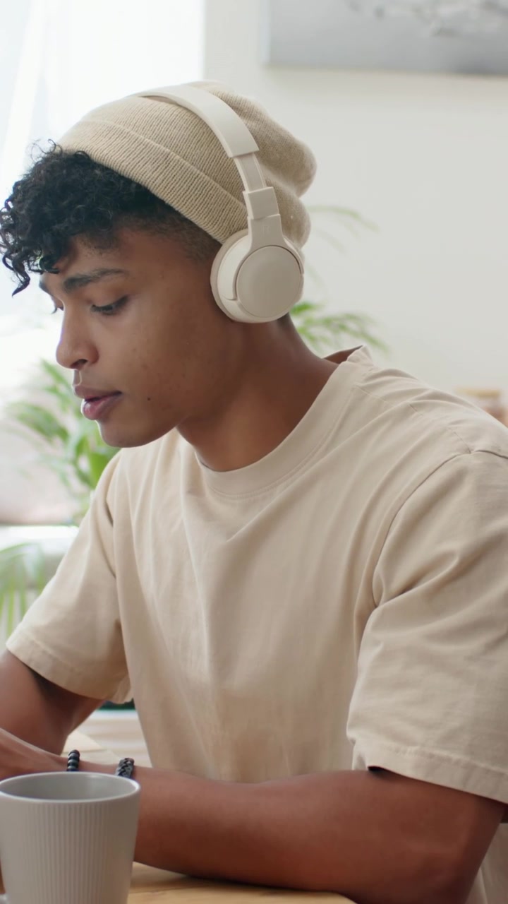 Vertical video: Sitting man in beanie and headset leaning at home table, gazing and reading device